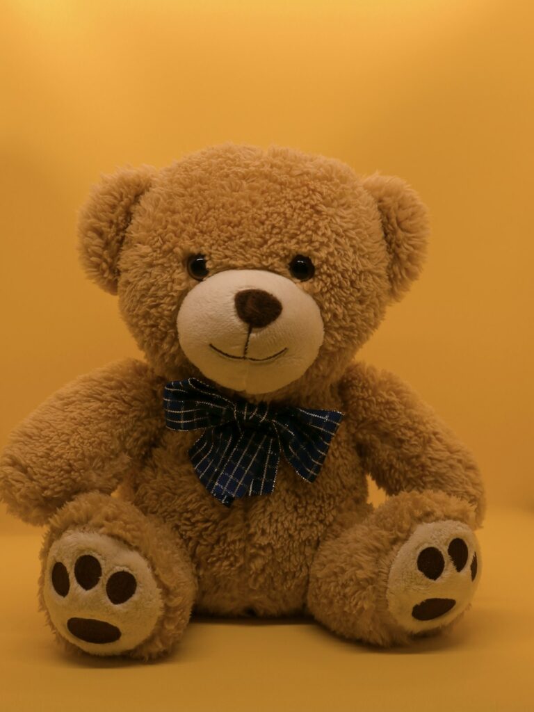 Soft tan plush teddy bear with a blue plaid bow tie on a warm yellow background, kept clean and cozy